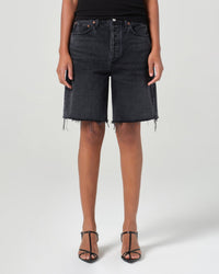 A person in a black sleeveless top, AGOLDE 90s Short in Hitch with frayed hems, and black heeled sandals stands against a plain light gray background.