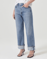 A person wears AGOLDE Fran Low Slung Straight in Invention jeans—light blue with a loose straight fit and wide cuffs—paired with a white top and black heeled sandals. The image is cropped at the shoulders against a plain background.