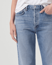 A person in AGOLDE Fran Low Slung Straight jeans in Invention, styled with a white t-shirt, stands with one hand in their pocket against a plain light background.