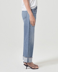 A person stands in profile against a white background, wearing a white T-shirt, AGOLDE Fran Low Slung Straight in Invention jeans with wide cuffs, and black heeled sandals.