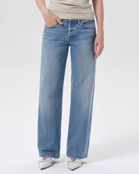 A person is shown from the waist down wearing AGOLDE’s Low Rise Loose in Vault jeans, styled with a beige top and white pointed shoes, standing against a plain light background.
