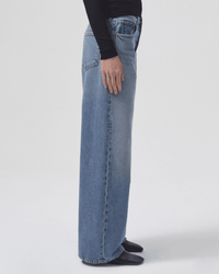 A person stands sideways against a plain white background, wearing AGOLDE’s Low Slung Baggy in Libertine light blue jeans with a high-waisted fit and wide legs, paired with a long-sleeve black top, hand resting by their side.
