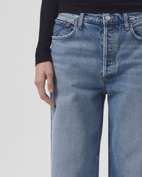 A person wears AGOLDE's Low Slung Baggy in Libertine jeans, high-waisted and light blue, paired with a long-sleeved black top. The focus is on the waist and upper legs, with one hand resting at the side.
