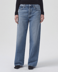 A person wears AGOLDE's Low Slung Baggy in Libertine—blue, high-waisted, relaxed-fit jeans with a slightly faded wash and wide straight legs in an organic cotton blend—and a black long-sleeve top, standing against a plain white background.