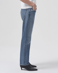 A person in profile wears the AGOLDE Parker Long in Invention jeans, a white shirt, and black heeled ankle boots. They stand against a plain white background with hands tucked casually in their pockets.