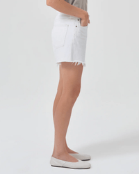 A person stands in profile against a white background wearing a light gray top, AGOLDE Parker Long Short in Panna Cotta with frayed hems, and white flats.