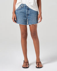 A person wears a white t-shirt, AGOLDE Parker Long Short in Ruin—high-rise, zero-stretch denim with frayed hems—and black sandals, standing against a plain light background. Image is cropped from the shoulders down.