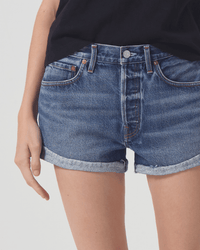 A person models the AGOLDE Parker Short in Ruin, high rise denim shorts with rolled cuffs, paired with a black shirt. Only their lower torso and upper legs are visible against a plain light background, left hand resting by their side.