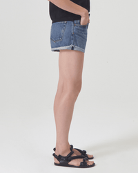 A person stands in profile against a plain white background, wearing AGOLDE's Parker Short in Ruin, a black top, and black braided-strap sandals.