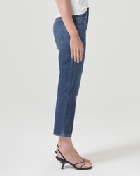 Someone stands sideways against a plain white background wearing AGOLDE’s Riley Crop in Control high-rise straight leg non-stretch jeans, paired with a white top and black heeled sandals.