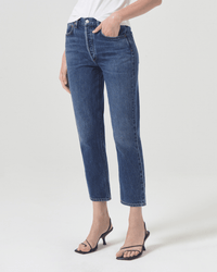 A person wearing AGOLDE's Riley Crop in Control high-rise straight leg jeans, a white top, and black strappy heeled sandals stands against a plain white background with one hand in their pocket and face not visible.