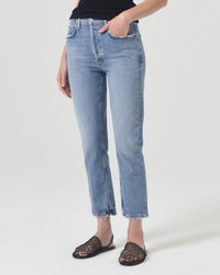 A person wears the AGOLDE Riley Crop in Quiver jeans, a black sleeveless top, and black woven sandals, standing against a plain white background with their left hand in their pocket.