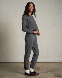 A woman stands sideways, smiling, wearing the Alexi French Terry Sweatshirt in Charcoal by perfectwhitetee with matching jogger pants, white socks, and burgundy loafers against a plain light-colored wall.
