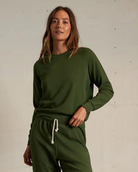 Wearing a matching Alexi French Terry Sweatshirt in Fern Green by perfectwhitetee, a person stands against a light wall with one hand on their hip and a relaxed expression.