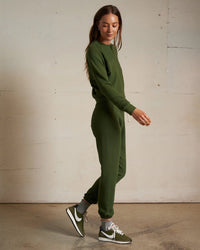 A woman in a perfectwhitetee Alexi French Terry Sweatshirt in Fern Green and matching joggers with green and white Nike sneakers stands sideways, looking down, against a plain light wall and concrete floor.