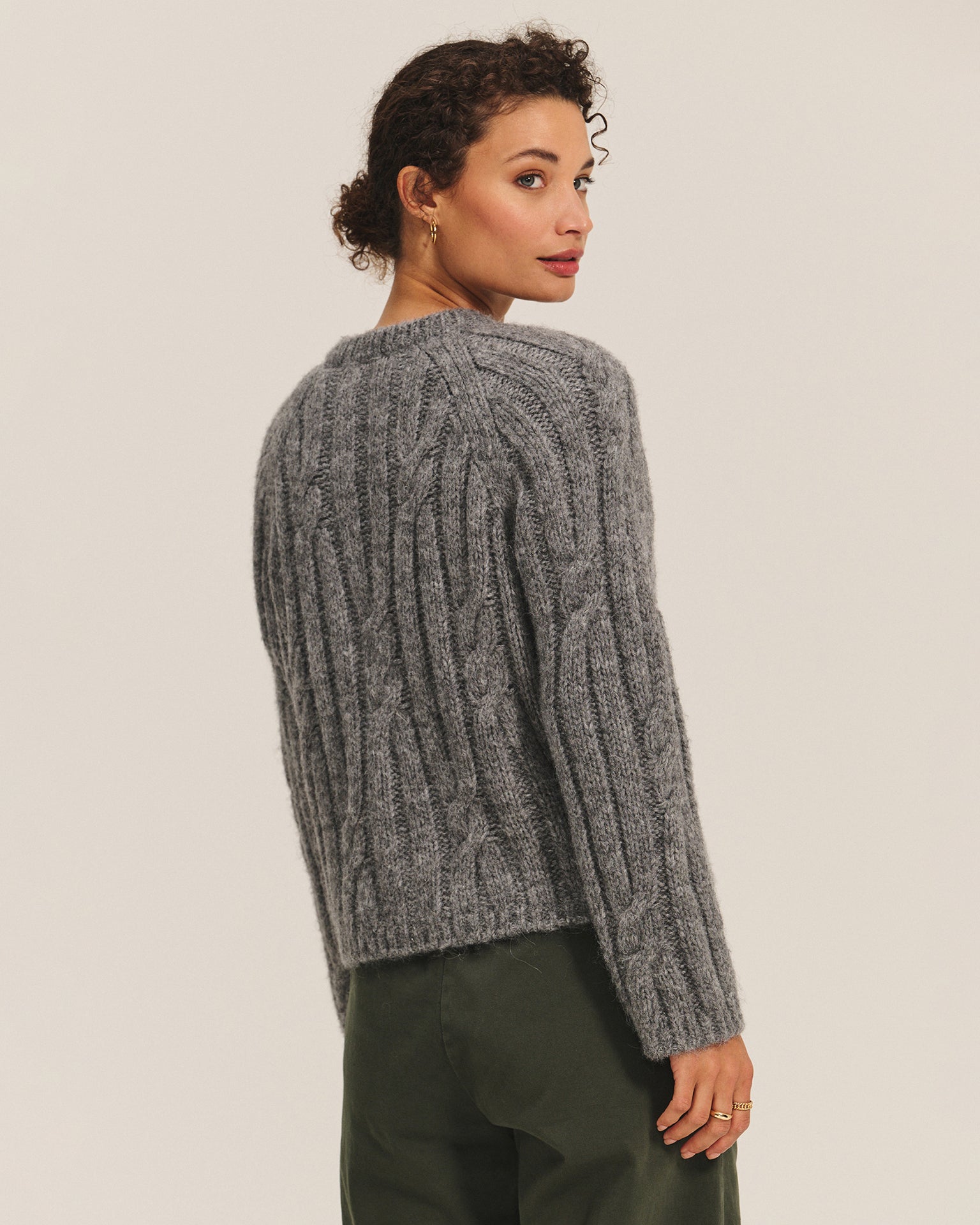 A woman with curly hair models the Velvet by Graham & Spencer Allison Long Sleeve Crew Cardi in Heather Grey, paired with green pants. She stands slightly turned to the side, gazing over her shoulder against a plain light background.