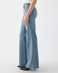 A person stands sideways against a plain white background, wearing AMO’s Anita Flare in Eternal—high-waisted, flared blue jeans made from comfort stretch denim—and black heeled boots.