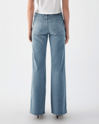 A person stands with their back to the camera, wearing AMO’s Anita Flare in Eternal—light blue comfort stretch denim jeans with a straight-leg cut, seam detail, two back pockets, and paired with heeled shoes and a white top.