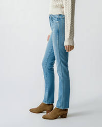 A person wears AMO’s Chloe Long Length in Liason light blue jeans, a cream cable-knit sweater, and tan suede ankle boots while standing against a plain light background. The image is cropped at the shoulders.