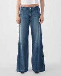A person in a white crop top and AMO's Edith Wide Leg Trouser in Romance stands against a plain light background. The denim features a faded wash, loose fit, and wide legs that cover the shoes.