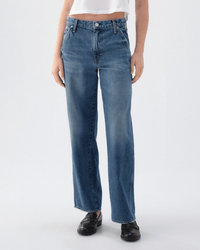 A person in AMO's Jocelyn Shaped Trouser in Two Timing, a women's mid rise straight leg jean, wears a white cropped top and black loafers, standing against a plain light background; photo is cropped at the shoulders.