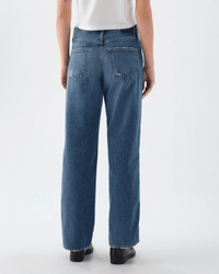 Facing away, the person wears a white shirt, black shoes, and AMO's Jocelyn Shaped Trouser in Two Timing—blue women's jeans with two back pockets and a relaxed fit—set against a plain light background.