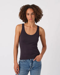 A person with curly hair models the AMO Layering Tank in Vintage Black with blue jeans, standing against a white background and looking at the camera with a relaxed expression, one hand in their pocket.