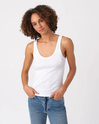 A person with curly hair wears the AMO Layering Tank in White and blue jeans, standing against a light background with one hand in their pocket and a relaxed expression.