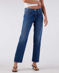 A person stands against a plain background wearing AMO’s Libby in Mastermind indigo wash, straight-leg jeans, paired with a white crop top and open-toe heels, one arm bent and the other relaxed.