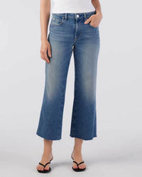 A person wears AMO's Marjorie in Dropout high-waisted, wide leg jeans with a cropped hem, paired with a white top and black sandals, standing against a plain light gray background.