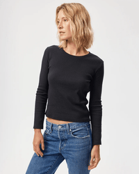 A woman with wavy blonde hair models the AMO Slim Rib Tee in Black with high-waisted blue jeans, standing against a plain white background and looking to her left.