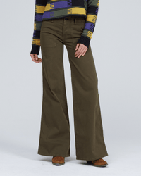 A person wearing ASKK NY's Boma Jean in Safari Green, a colorful patterned sweater, and brown shoes stands against a plain light gray background.