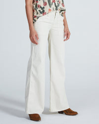 A person wearing high-waisted, wide-leg ASKK NY Boma Twill Jeans in Sand, a cactus-patterned sleeveless top, and brown shoes stands against a plain background.
