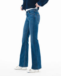 Wearing ASKK NY Bond Street in Paris blue mid-rise bootcut jeans in Japanese stretch denim, a dark blue sweater, and white pointed-toe shoes, standing against a white background. Image is cropped above the shoulders.