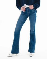 Wearing the ASKK NY Bond Street in Paris mid-rise, dark blue flare jeans made from Japanese stretch denim, a person stands against a white background with their hands near the waistband, paired with a navy sweater and white heeled shoes.