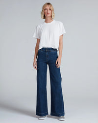 A person stands facing forward against a light gray background, wearing a plain white t-shirt, ASKK NY Brighton in Dark Stone high rise wide leg jeans made from Japanese stretch denim, and striped slip-on shoes.