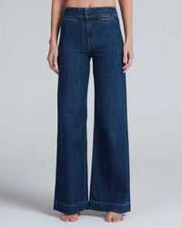A person wearing ASKK NY's Brighton in Dark Stone high rise, wide leg jeans stands barefoot against a light gray background. Made from Japanese stretch denim, the jeans feature front pockets and a double button closure.
