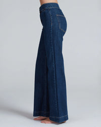 A person stands barefoot in ASKK NY's Brighton in Dark Stone high rise wide leg jeans made from Japanese stretch denim. Photo is side view, showing the jeans’ length and fit; upper body is not visible, background is plain and light.