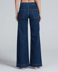 A person is shown from the back, barefoot against a plain light background, wearing ASKK NY's Brighton in Dark Stone jeans. This high-waisted style has large back pockets, visible stitching, and is crafted from Japanese stretch denim.