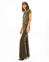 A woman with long brown hair stands against a white background, wearing an olive green t-shirt and the ASKK NY Brighton Corduroy Wide Leg pant in Safari Green, paired with black heeled shoes. She looks slightly to the side.