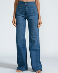 A person stands barefoot against a plain light gray background, wearing the ASKK NY Brighton Wide Leg in Mercury—high-waisted jeans with front pockets and visible orange stitching.