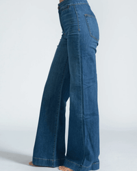 A person stands barefoot in ASKK NY Brighton Wide Leg jeans in Mercury, made from blue Japanese stretch denim, photographed from the side against a plain light background.