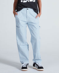 Wearing ASKK NY’s Carpenter in Blue Stripe pants, a black graphic tee, and black-and-white sneakers, this person stands with hands in pockets against a plain background.
