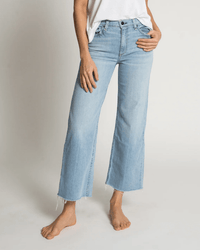 A person stands barefoot against a plain background, wearing light blue ASKK NY Crop Wide Leg in Zuma jeans with raw hems and a white t-shirt.