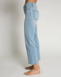 A person stands barefoot on a light floor, shown in side view from waist to feet, wearing ASKK NY’s Crop Wide Leg in Zuma—high-waisted, light blue jeans with frayed hems—against a plain background.