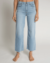 A person wearing ASKK NY's Crop Wide Leg in Zuma—light blue, wide-leg jeans with a raw hem—stands barefoot against a plain white background, showcasing this perfect summer denim style.