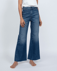 A person models the Cropped Brighton in Daytona jeans by ASKK NY—a pair of high rise, wide leg blue jeans in Japanese stretch denim with raw hems—wearing a white shirt and standing barefoot against a plain light background.