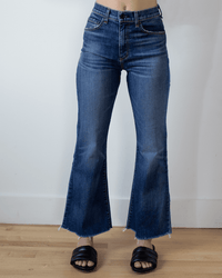 A person in ASKK NY’s Geek in Auburn high rise crop flare jeans with raw hems and black open-toed slide sandals stands on a wooden floor against a white wall.