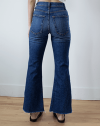 A person wearing ASKK NY's Geek in Auburn dark blue jeans with a raw hem and black sandals stands on a wooden floor against a plain white wall, viewed from behind.