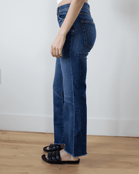 A person stands indoors on a wooden floor, seen from the side, wearing ASKK NY's Geek in Auburn high rise crop flare jeans with a raw hem and black slide sandals.